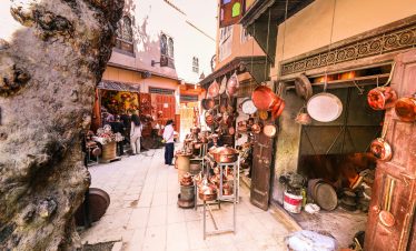 A vibrant outdoor market scene displaying hanging brass pots and people shopping.