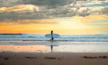 A serene view of a surfer walking along the beach with a surfboard at sunset in Essaouira, Morocco.